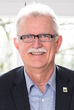 Ein Mann mit weißen Haaren und dunkler Brille steht vor einem Fenster und lächelt